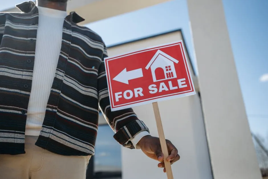 Photo by Pavel Danilyuk on Pexels Close-up of a person holding a for sale sign outside a modern house.