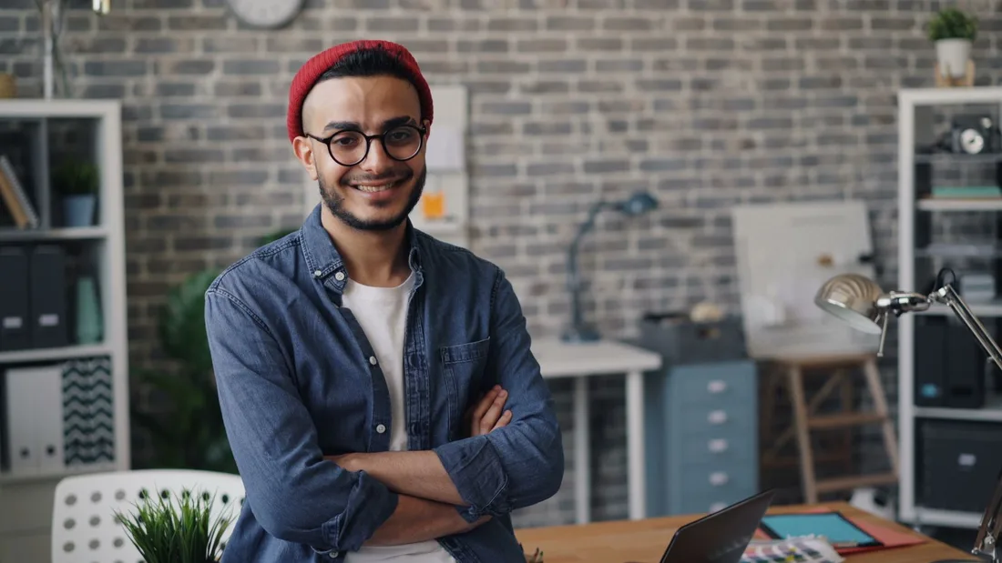 Smiling young man standing confidently in a modern office with a brick wall background in the UK. Photo by Vitaly Gariev on Pexels
