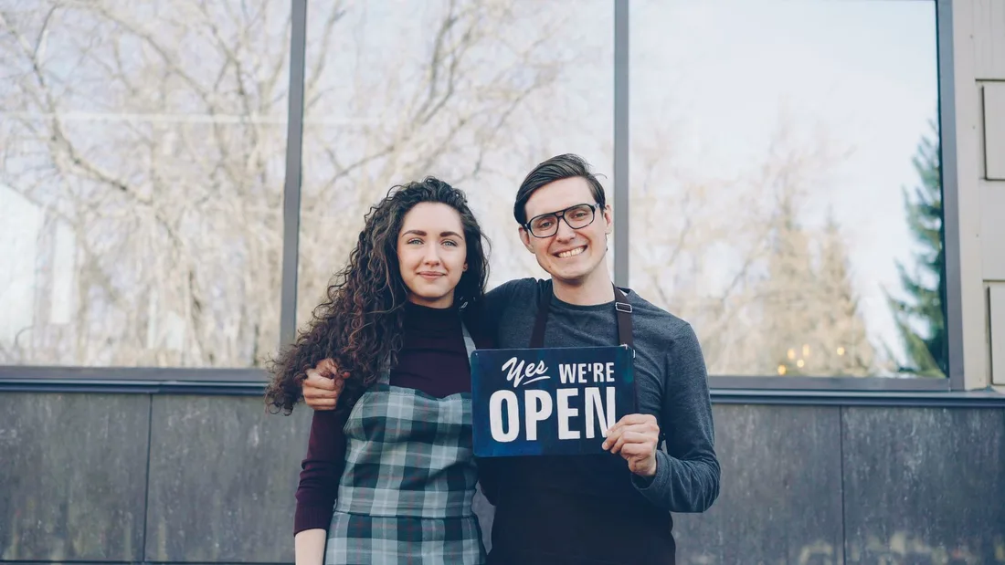 Two small-business owners welcoming customers with an open sign. Photo by Vitaly Gariev on Pexels